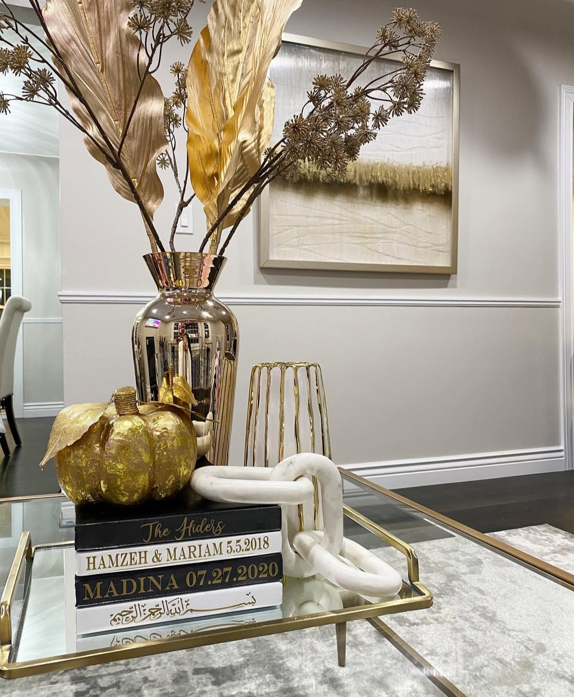Glam glass coffee table featuring a metallic gold vase with large gold leaves, marble chain on personalized books, and a gilded pumpkin.