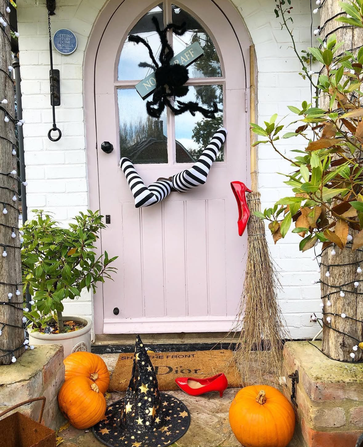 Pale pink arched door with a huge fuzzy spider, striped witch legs, broom, pumpkins, and a starry witch hat; one red heel on the step.