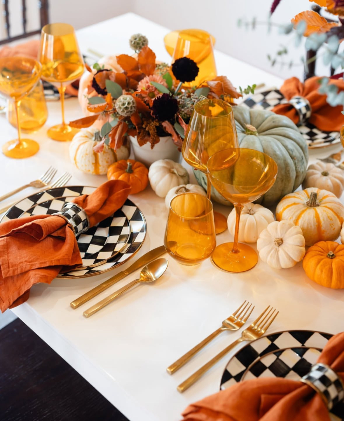 Bold autumn table with amber stemware, checkered plates, orange napkins, and piled pumpkins.