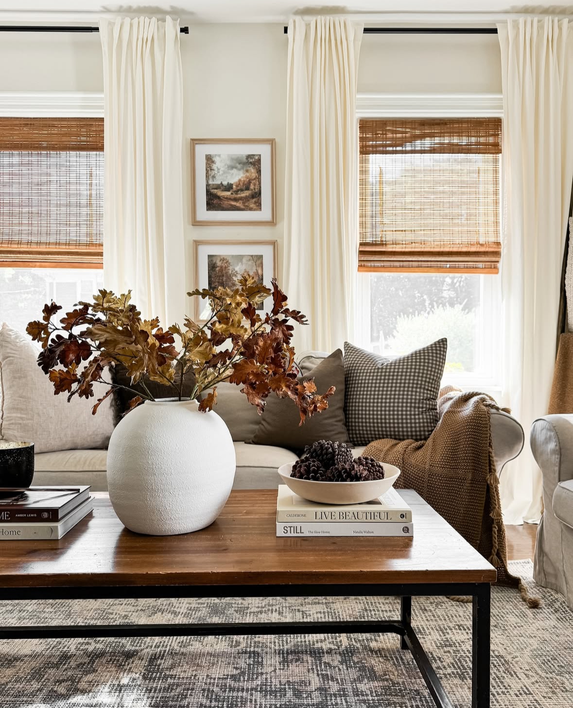 Long coffee table styled with a round white vase of copper leaves, a bowl of pinecones on design books, and neutral pillows with gingham accents.