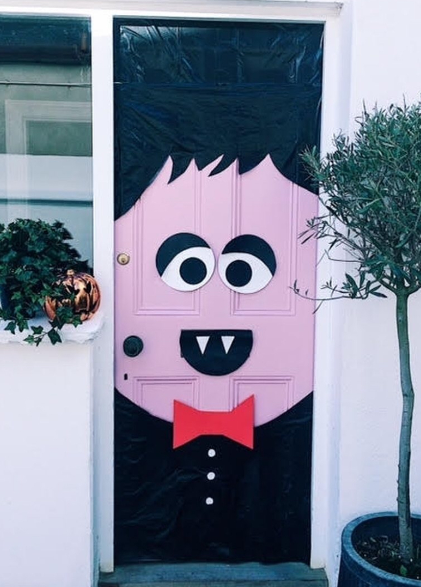 Pink door transformed into a cute vampire with black hair, fanged mouth, and red bow tie; potted plants on either side.