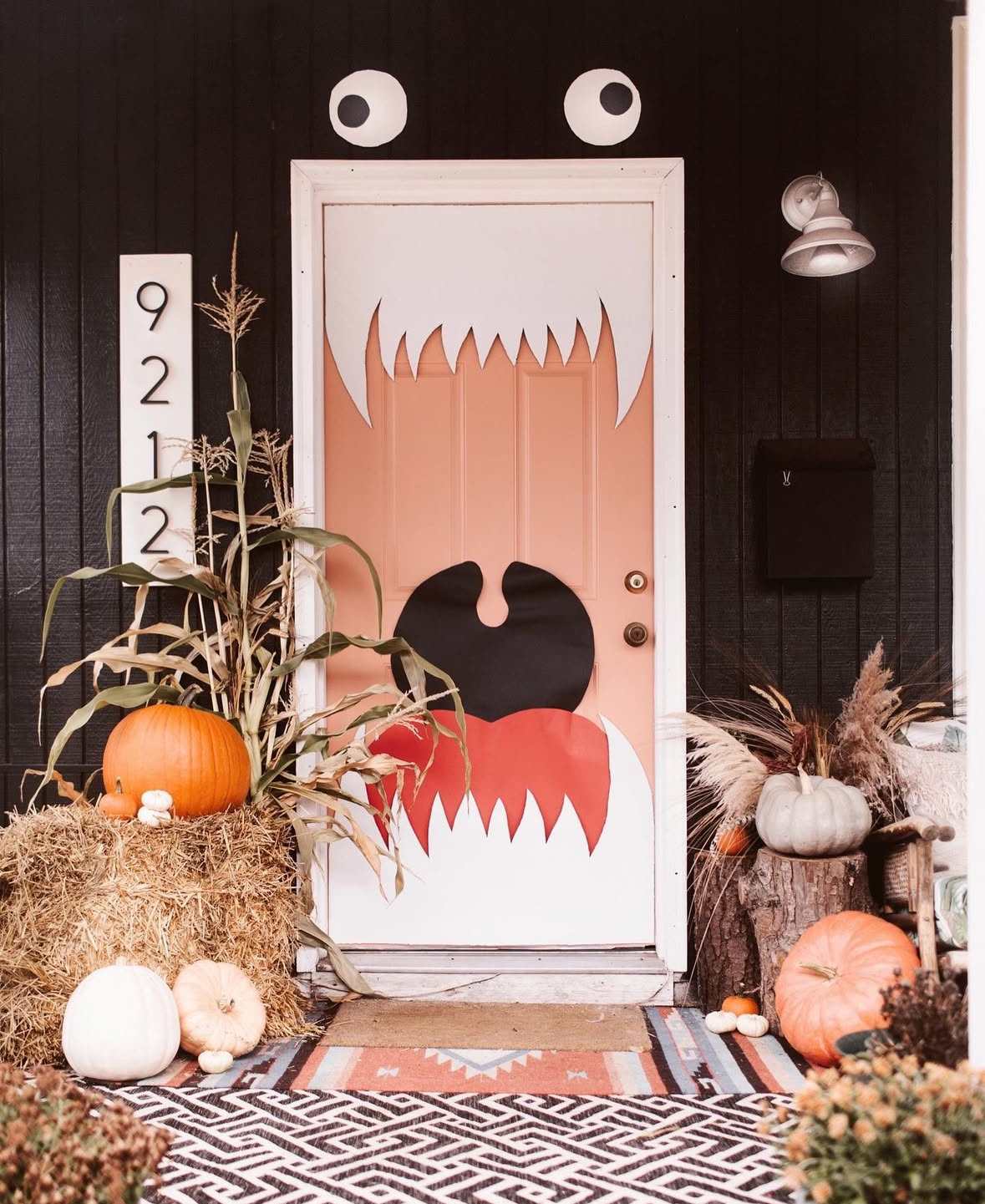 Coral door made into a monster mouth with paper eyes and jagged white teeth; pumpkins, hay bale, and fall grasses on a patterned porch rug.