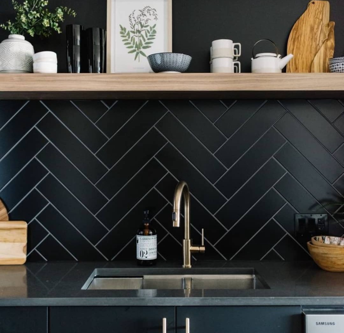Matte black herringbone tile backsplash with a wood shelf and brass faucet