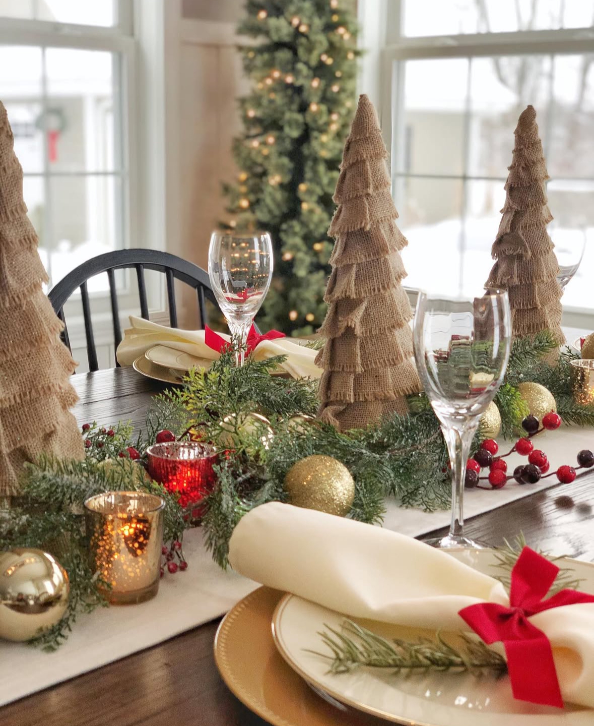 Burlap cone tree centerpieces with evergreen garland, red berries, gold ornaments, and cream napkins tied with red bows and rosemary sprigs.