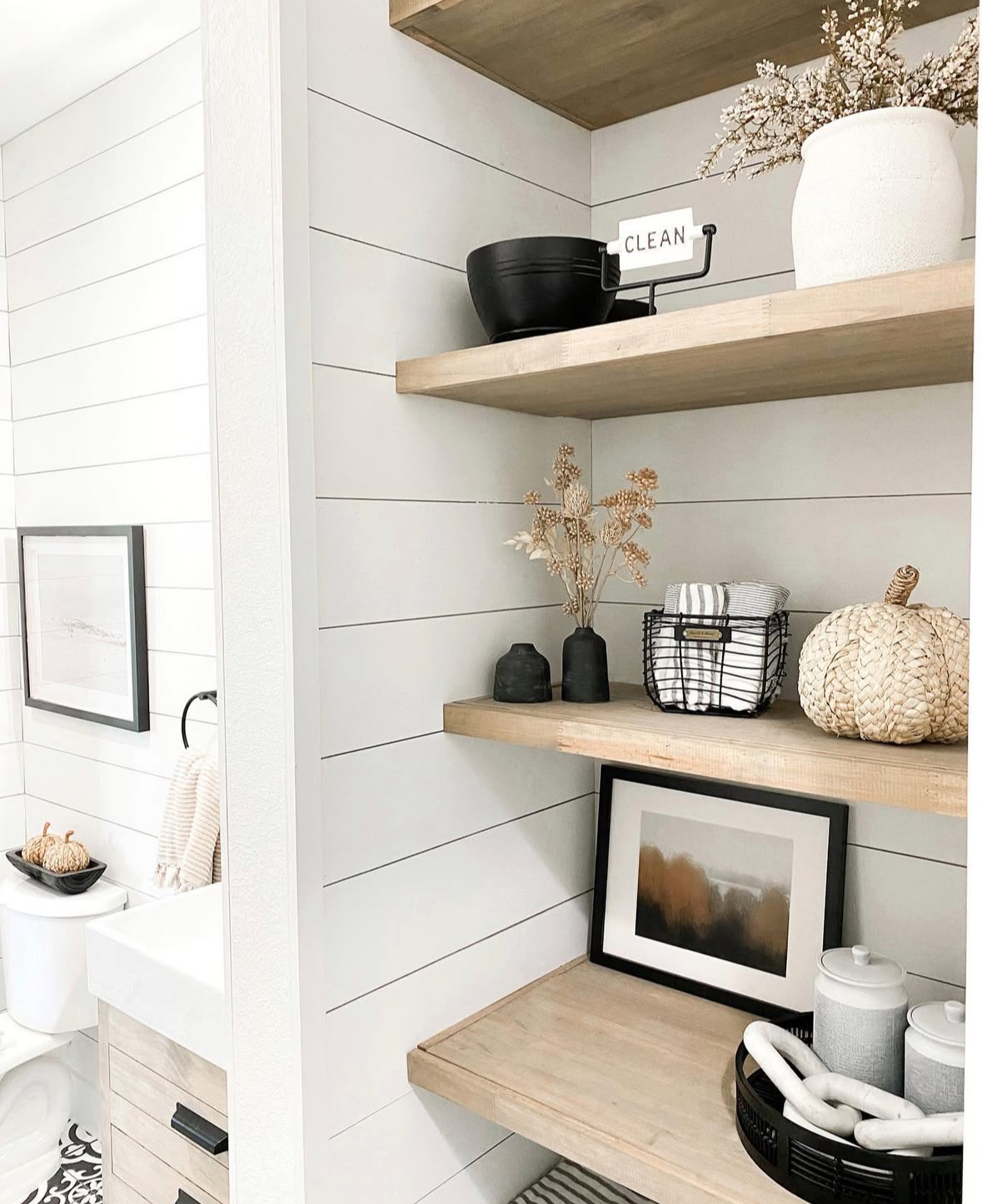 Bathroom corner shelves with shiplap, woven pumpkin, dried stems, black accents, and rolled towels.