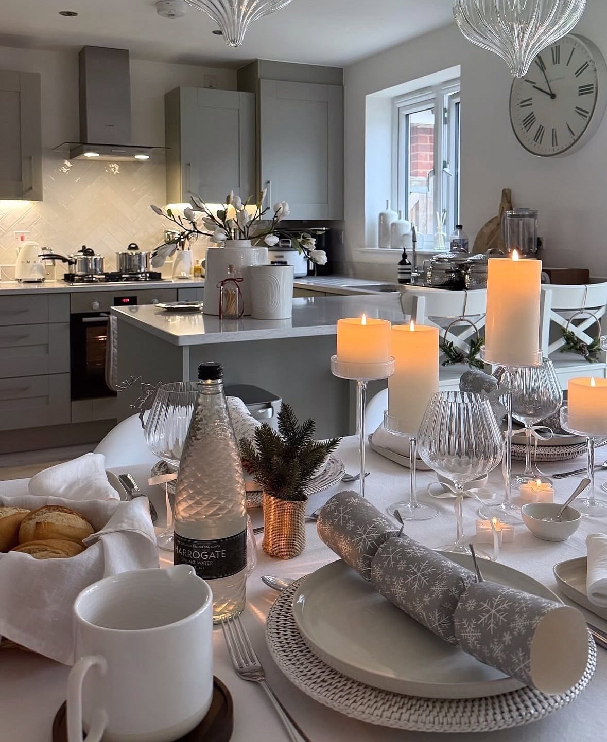 Neutral kitchen backdrop with a round table styled in all white: stacked plates, gray crackers, ribbed glassware, and clusters of glowing pillar candles.