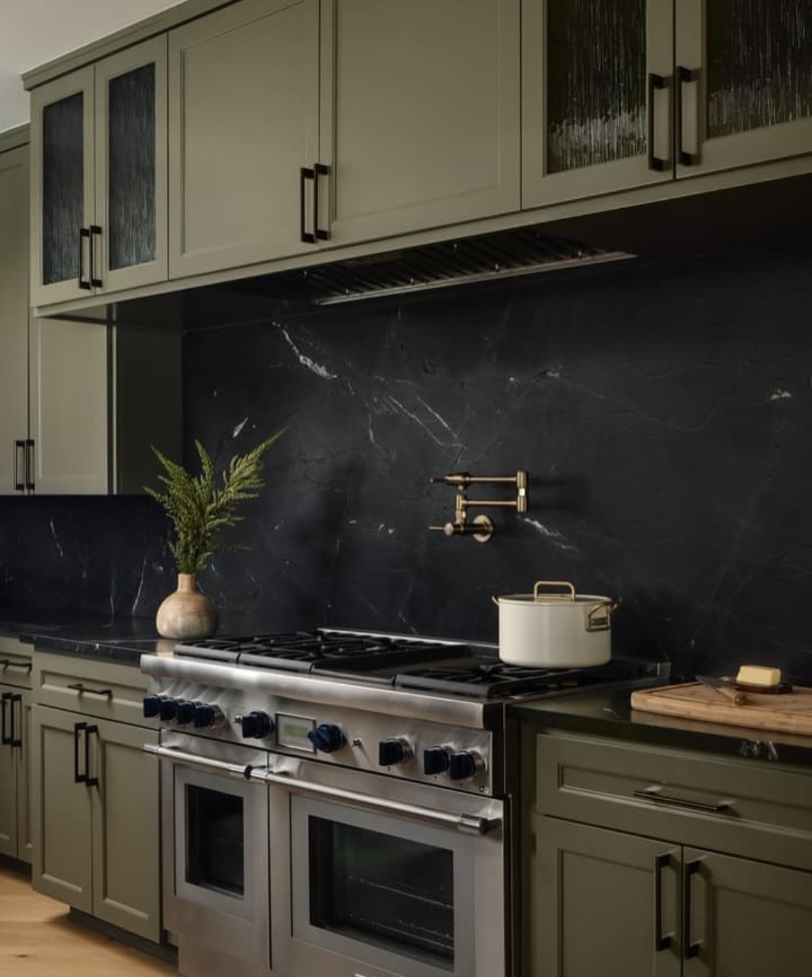 Olive kitchen cabinets paired with a black slab backsplash and brass pot filler
