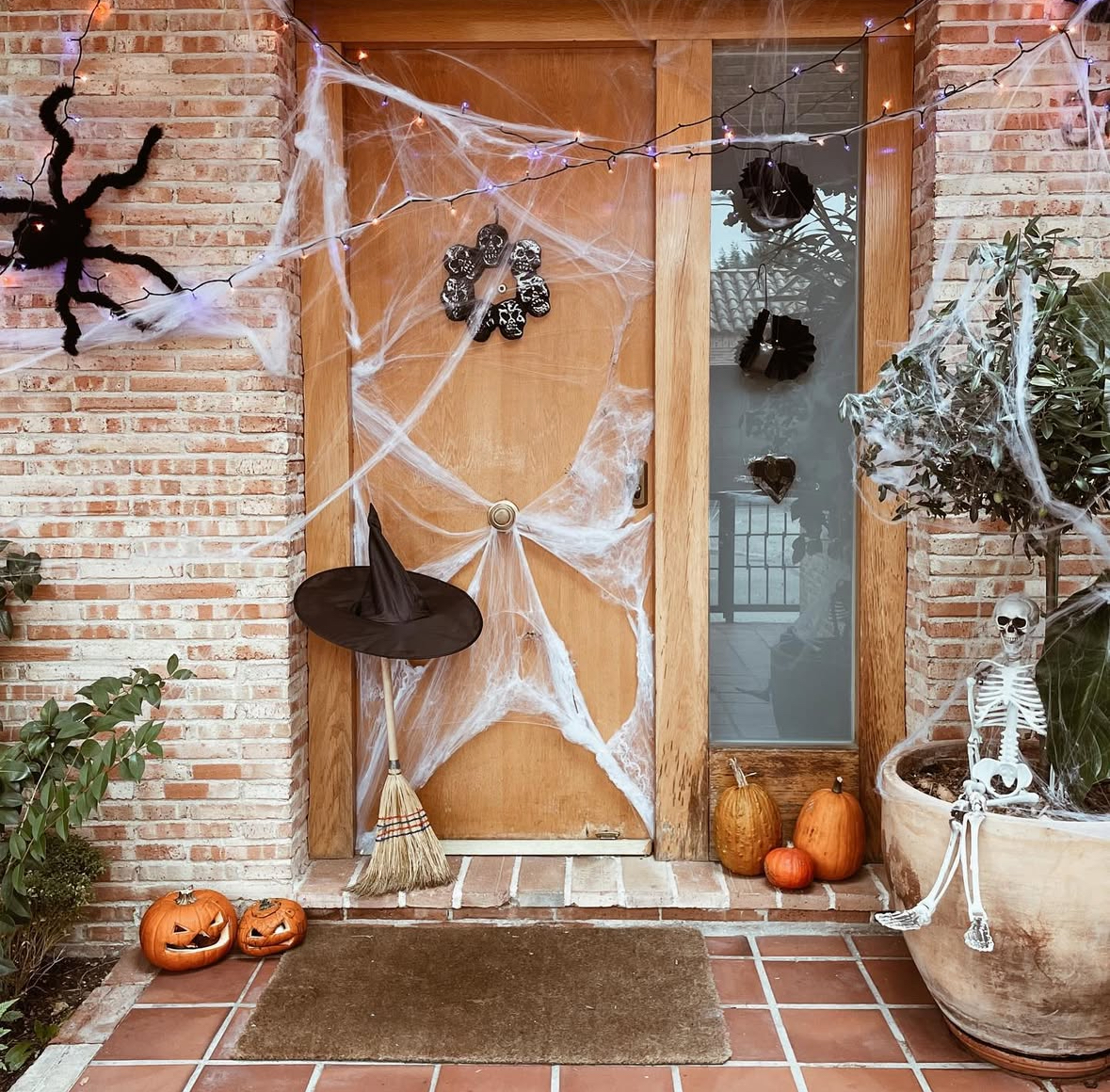 Natural wood door wrapped in faux spider webs with a giant spider, witch hat and broom, string lights, pumpkins, and a seated skeleton.