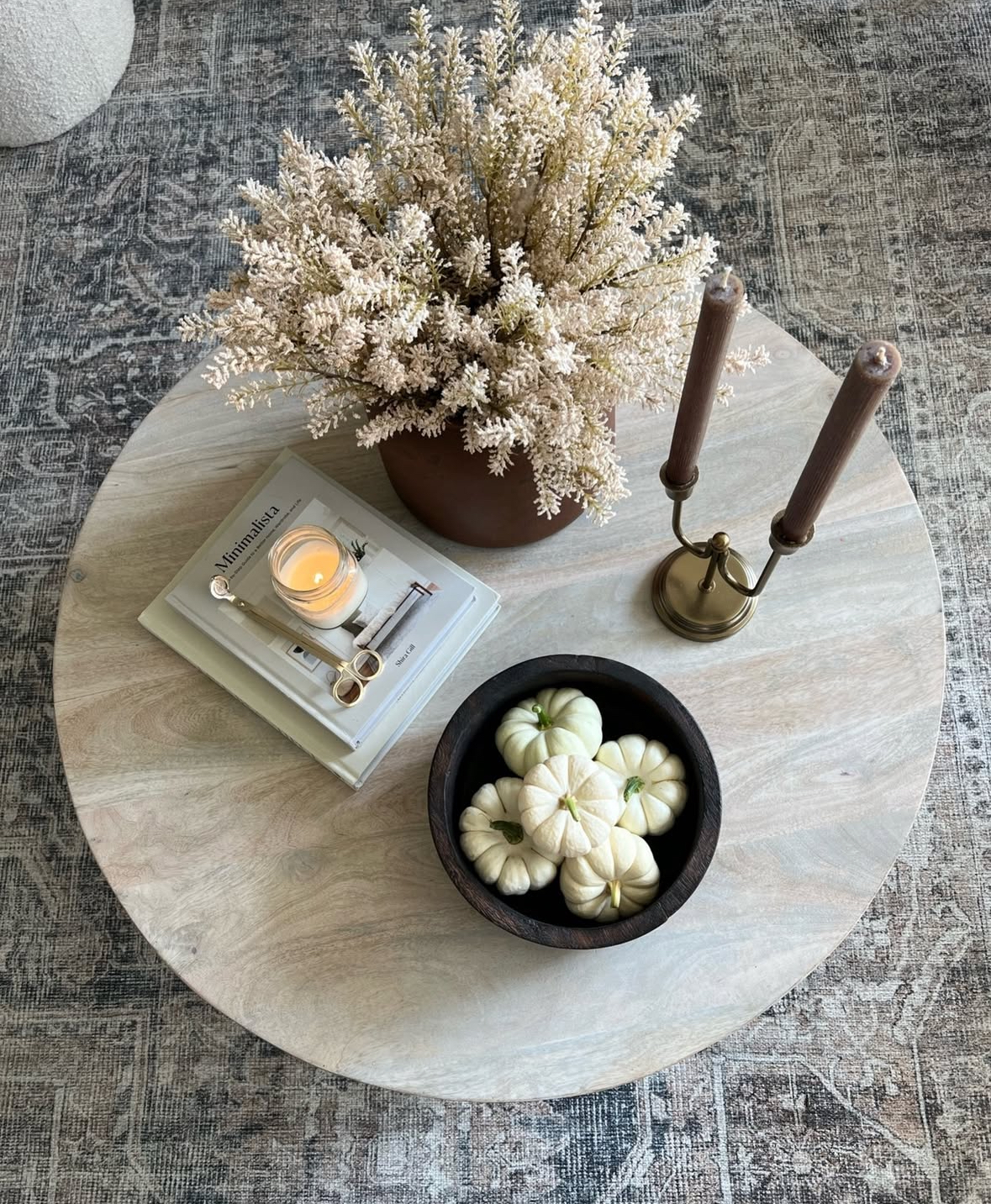 Close-up of round wood coffee table styled with a brown vase of pale foliage, candle on stacked books, brass candlestick, and bowl of white pumpkins.