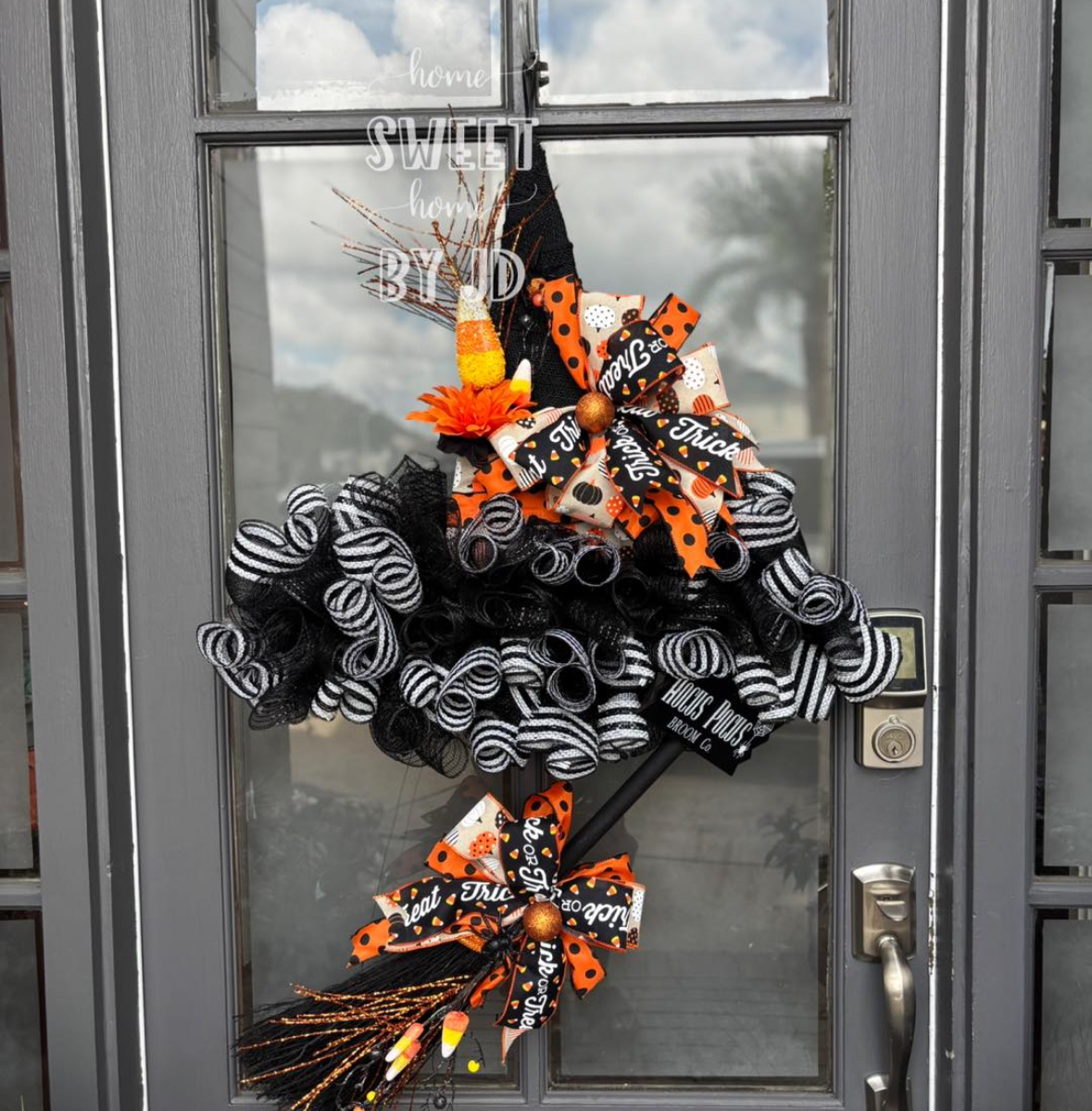 Witch-hat shaped wreath made from black mesh curls, striped ribbon, candy-corn picks, and a broom tail on a glass door.