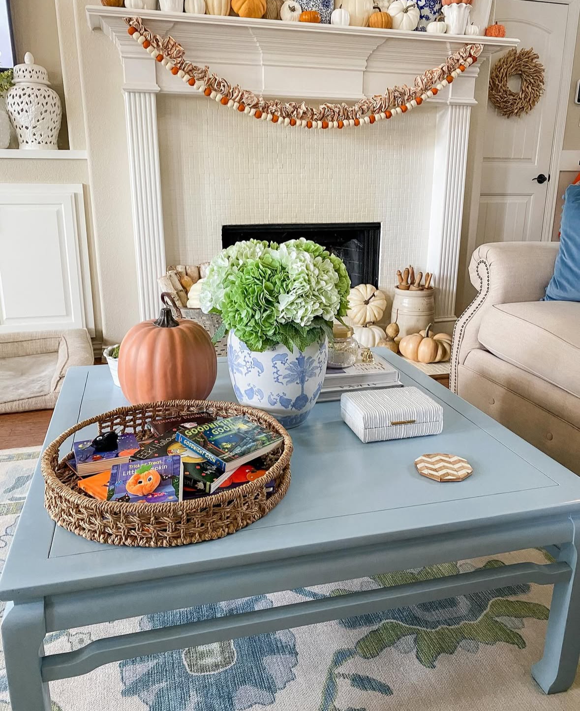 Blue coffee table with woven tray of children’s books, pumpkin decor, and a blue-and-white vase of hydrangeas; mantel lined with pumpkins behind.