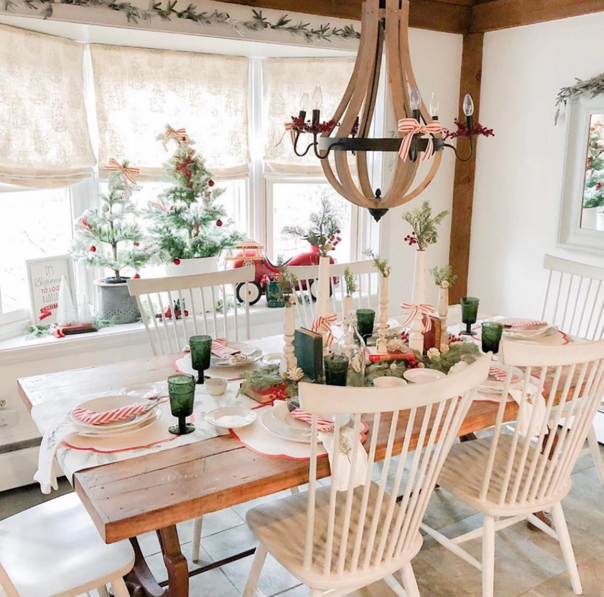 Farmhouse table with white spindle chairs, green goblets, mini trees, and candy-cane ribbons under a wooden chandelier; bright windows behind.