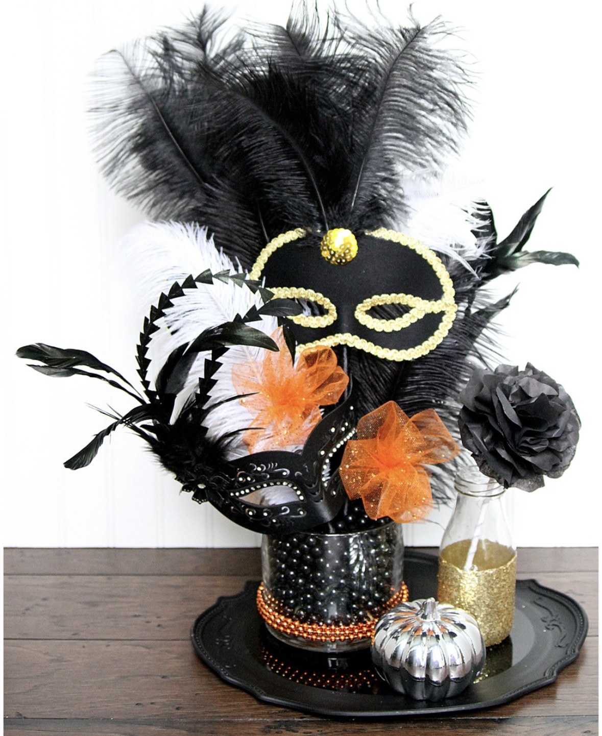 Black-and-gold Halloween centerpiece with feather plume, mini masquerade mask, beads, and metallic pumpkins on a tray