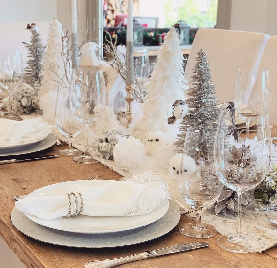 Snowy white centerpiece with glitter trees, faux owls, silver accents, and lace-edged napkins on a wood table.