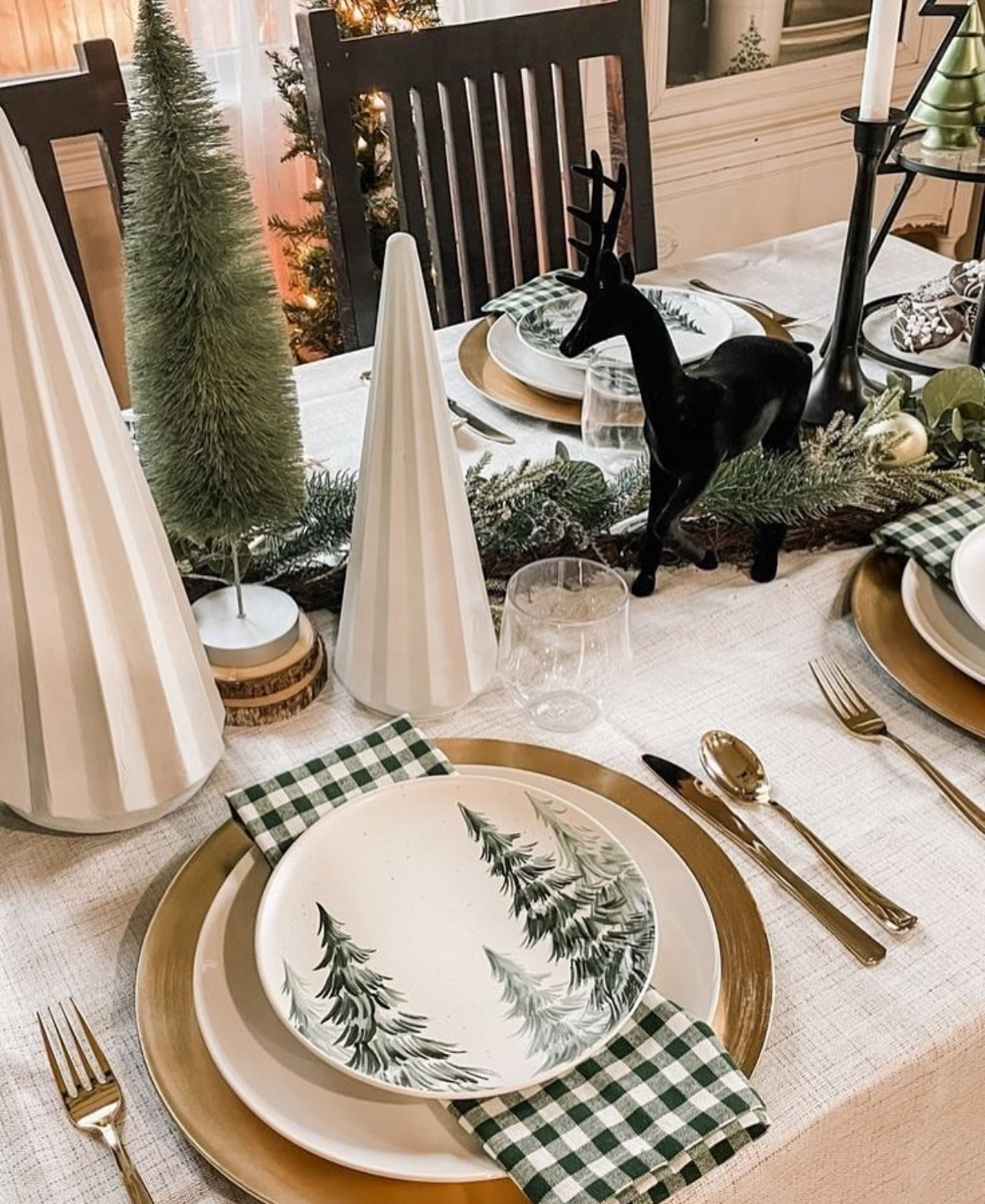 Neutral table with gold chargers, fir-tree plates, green gingham napkins, a black deer figurine, and cone trees.