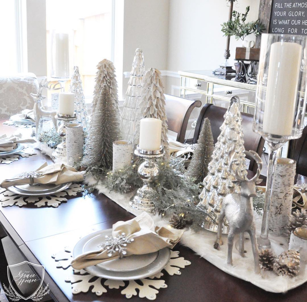 Winter-white centerpiece of mercury glass trees, faux snow, birch accents, candles, and silver deer with snowflake placemats on a dark wood table.