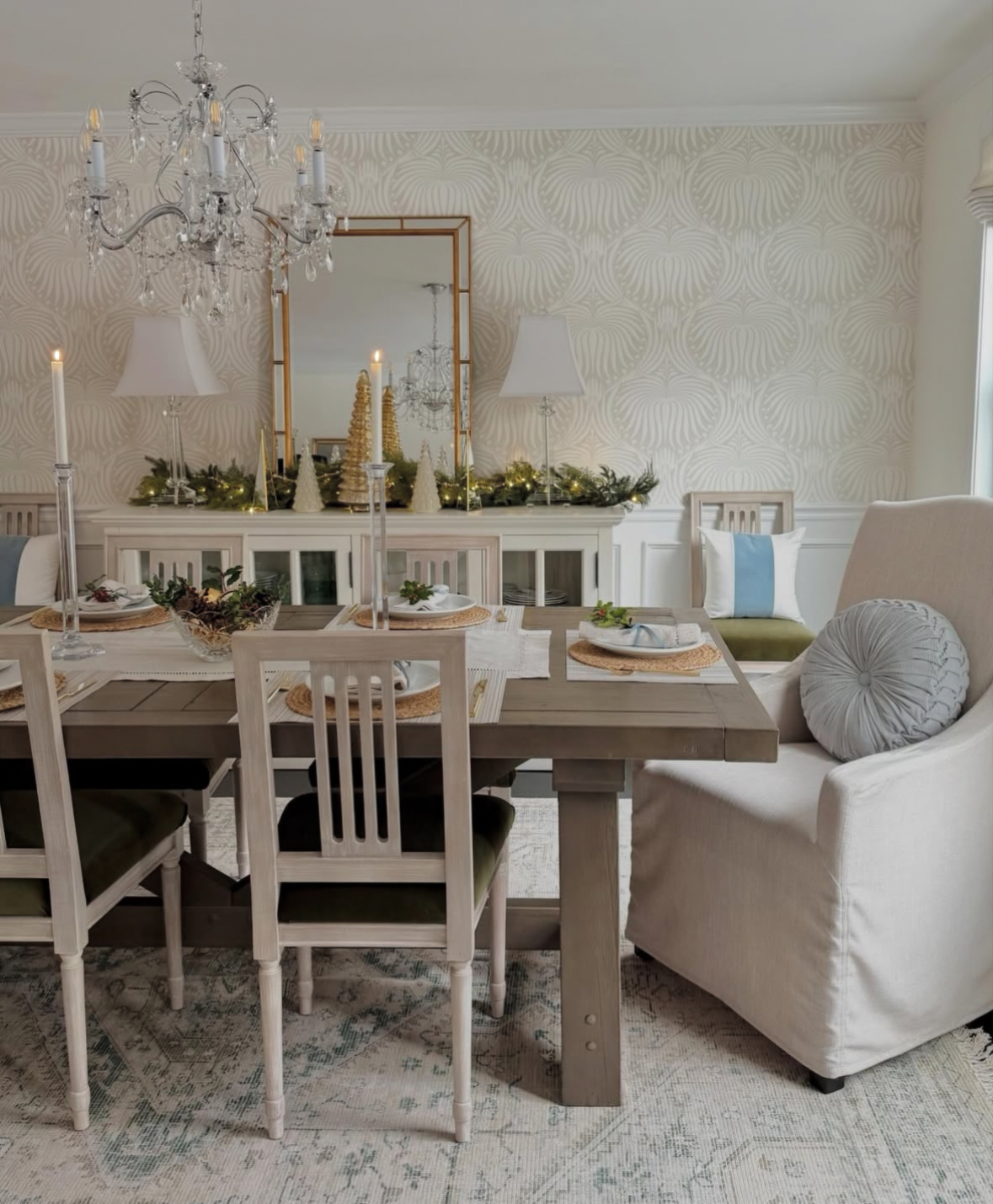 Neutral dining room featuring simple woven-charger place settings and a sideboard garland with bottle-brush trees, twinkle lights, and crystal candlesticks.