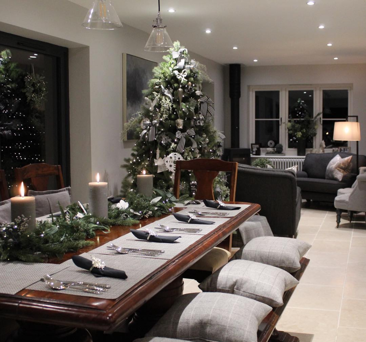 Moody gray tablescape with evergreen garland and chunky candles, gray napkins at each setting, and a black-and-white ribboned Christmas tree nearby.