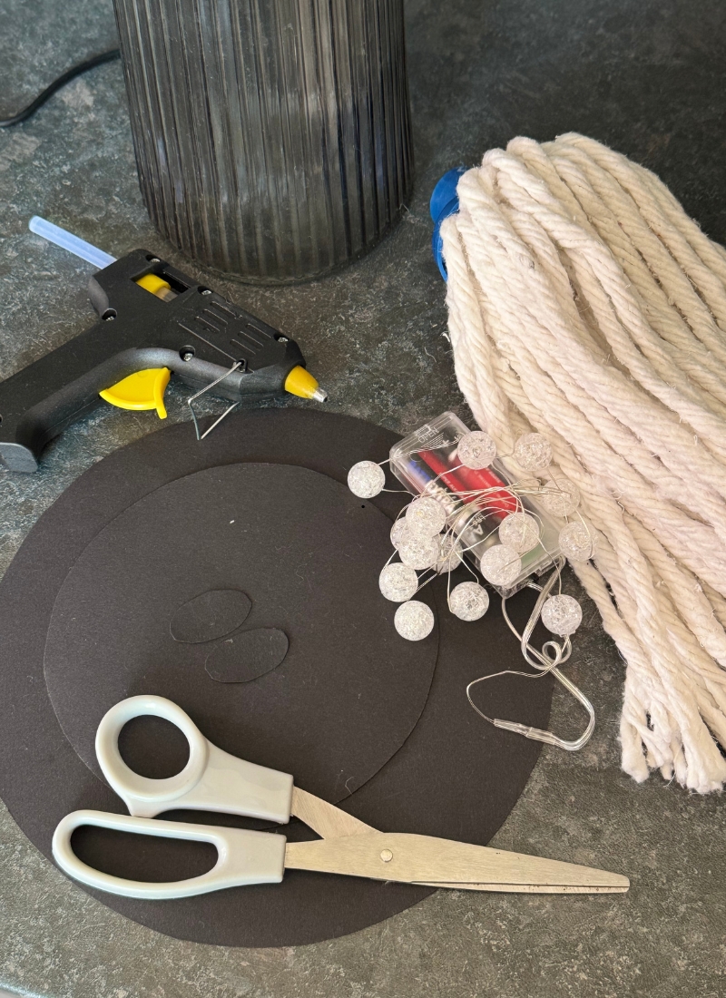 DIY Halloween ghost supplies: glass vase, cotton mop head, black paper, LED lights, scissors, and hot glue gun on counter.