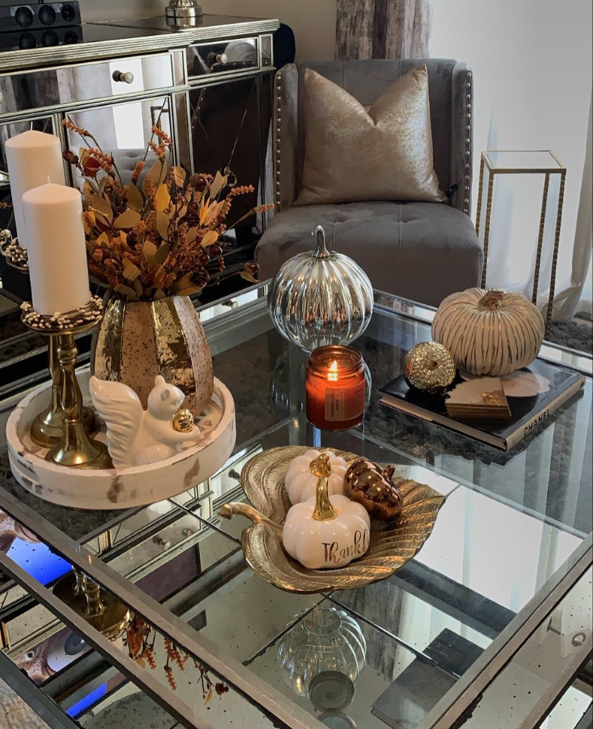 Mirrored coffee table arranged with marble tray, gold-flecked vase of fall stems, brass candlesticks, leaf dish with ceramic pumpkins, and a glowing candle.