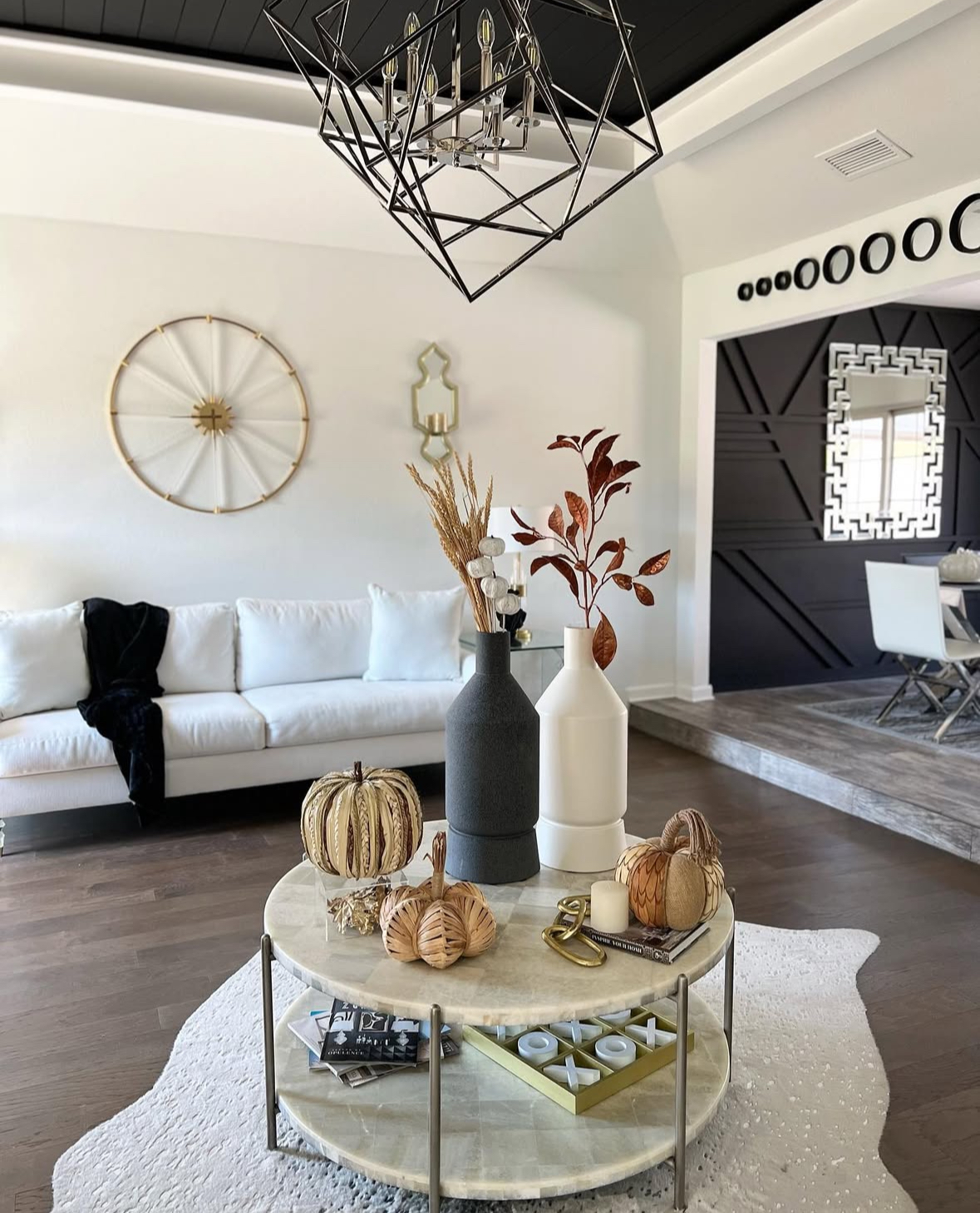 Modern living room with round marble two-tier coffee table decorated with tall sculptural vases, woven pumpkins, gold chain, and tic-tac-toe set.