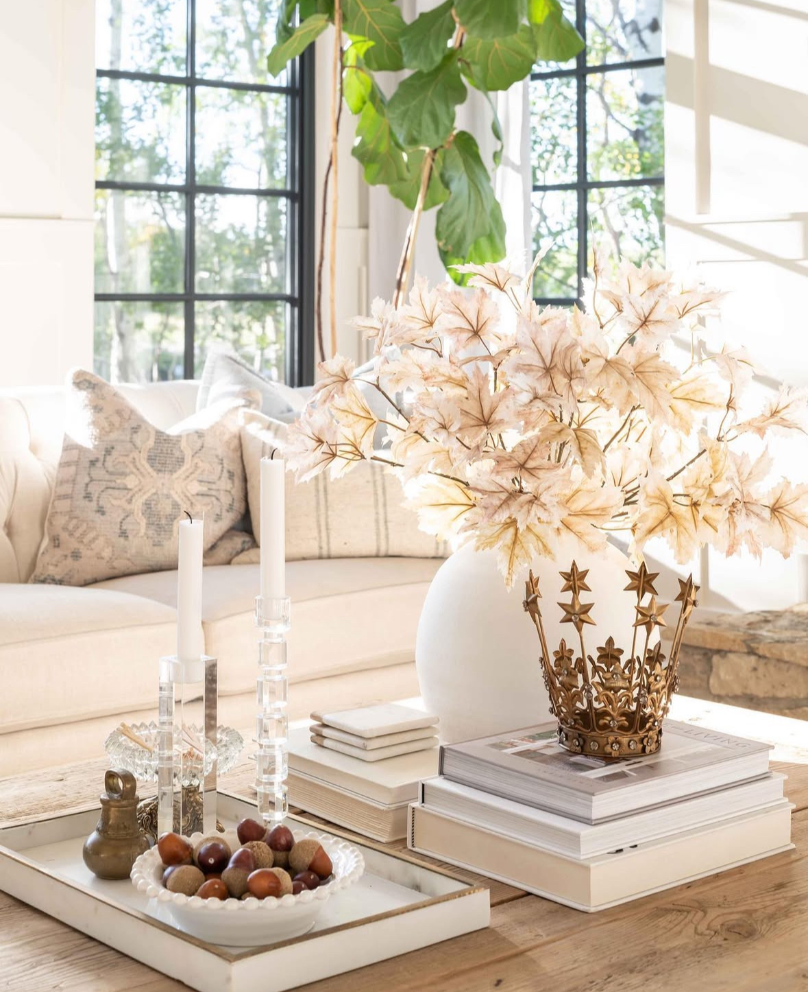 Sunlit neutral space with wood coffee table; oversized white vase of pale maple leaves, crystal tapers on a tray, nut bowl, stacked books, and a decorative crown.