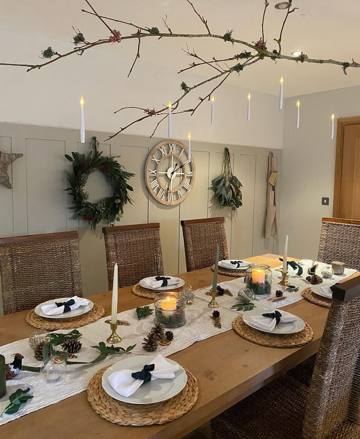 Foraged branch overhead with hanging LED tapers above a natural table setting of woven chargers, pinecones, greenery, and simple white plates.