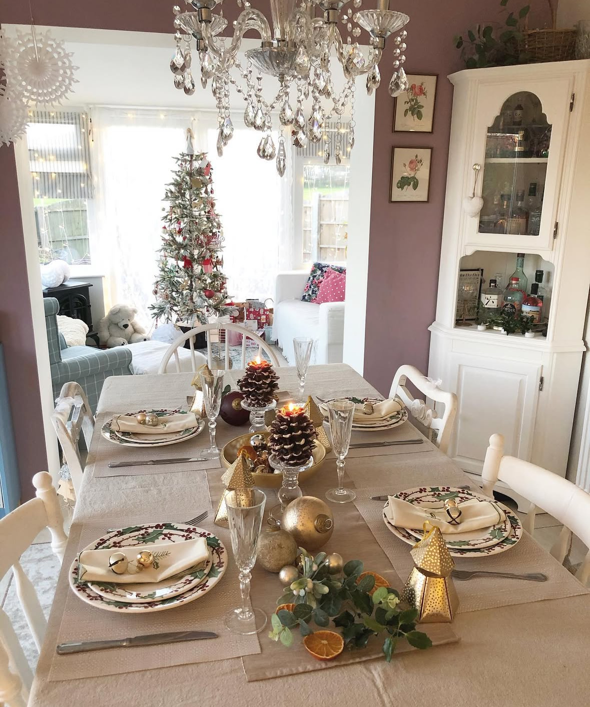 Cozy dining room table styled with holly plates, pinecone candles, dried orange slices, gold ornaments, and a crystal chandelier; small tree in the background.