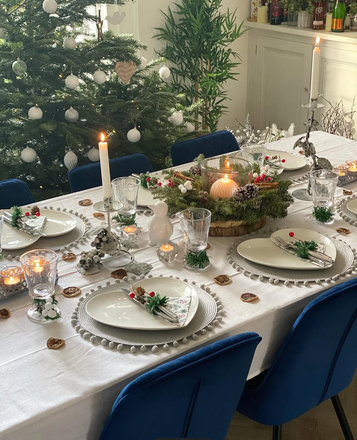 White tablecloth with pom-trim placemats, simple white plates, taper candles, and a wreath centerpiece; blue chairs and a white-ornamented tree nearby.