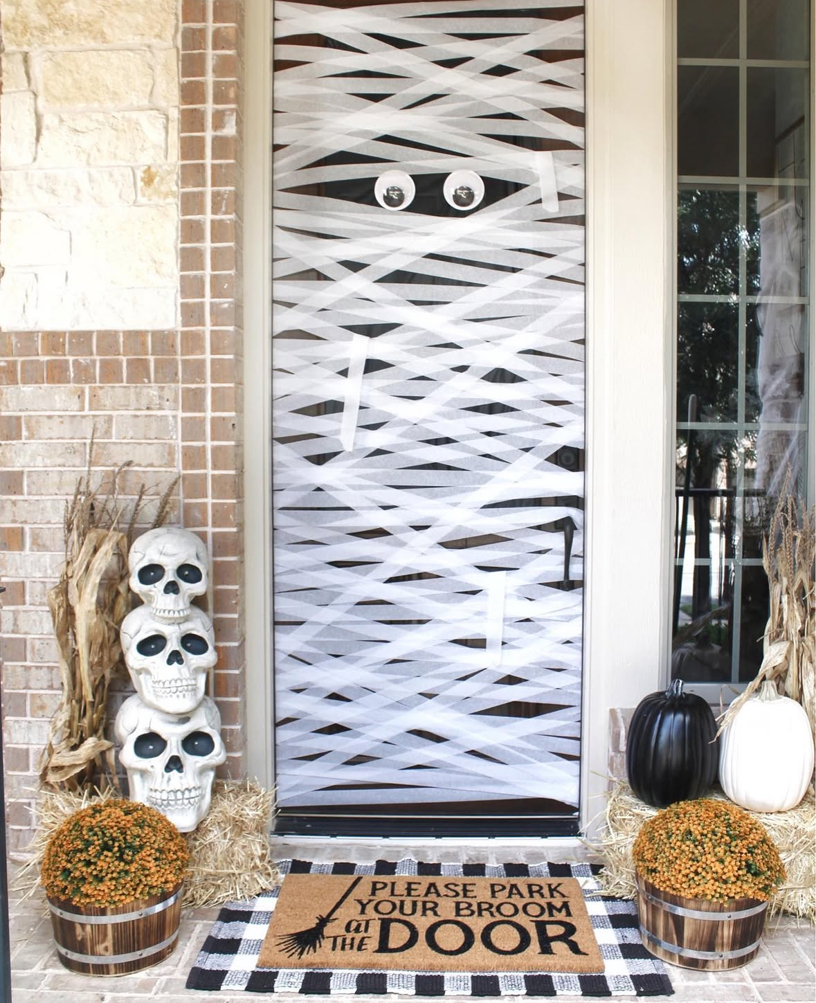 Screen door wrapped like a mummy with white streamers and big eyes; stacked skulls, pumpkins, hay bales, and a “Please park your broom at the door” mat.