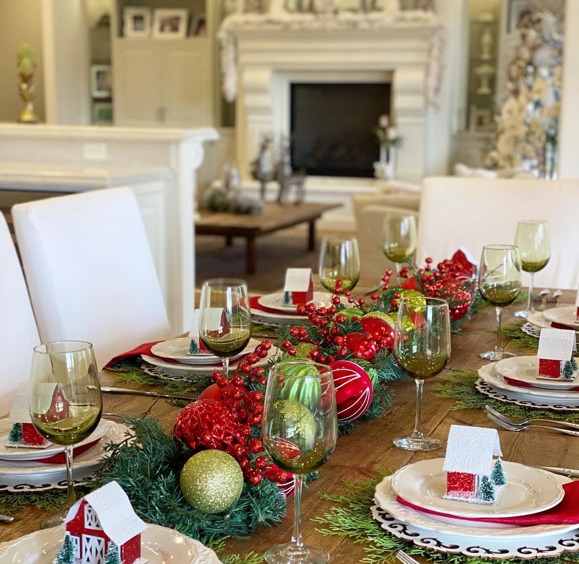 Long wood table with a runner of red berries and oversized ornaments, olive wine glasses, and white plates topped with tiny house place cards.