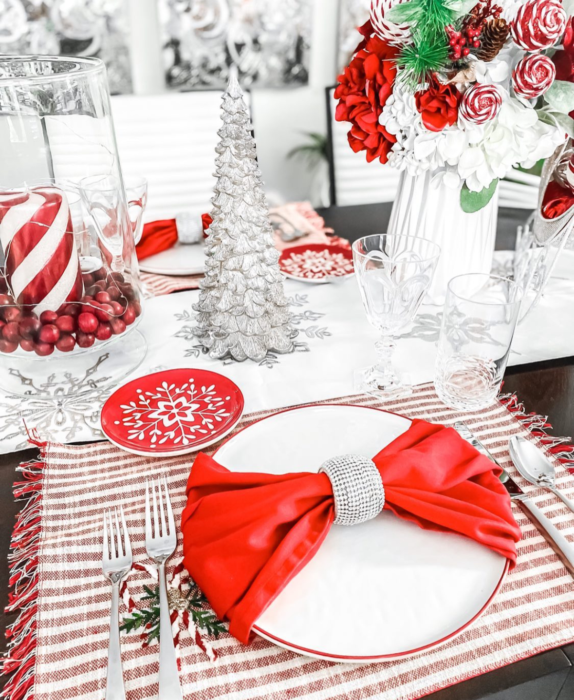 Close-up place setting with a rhinestone-cuffed red bow napkin on white china, striped placemat, silver tree décor, and a glass hurricane filled with cranberries.