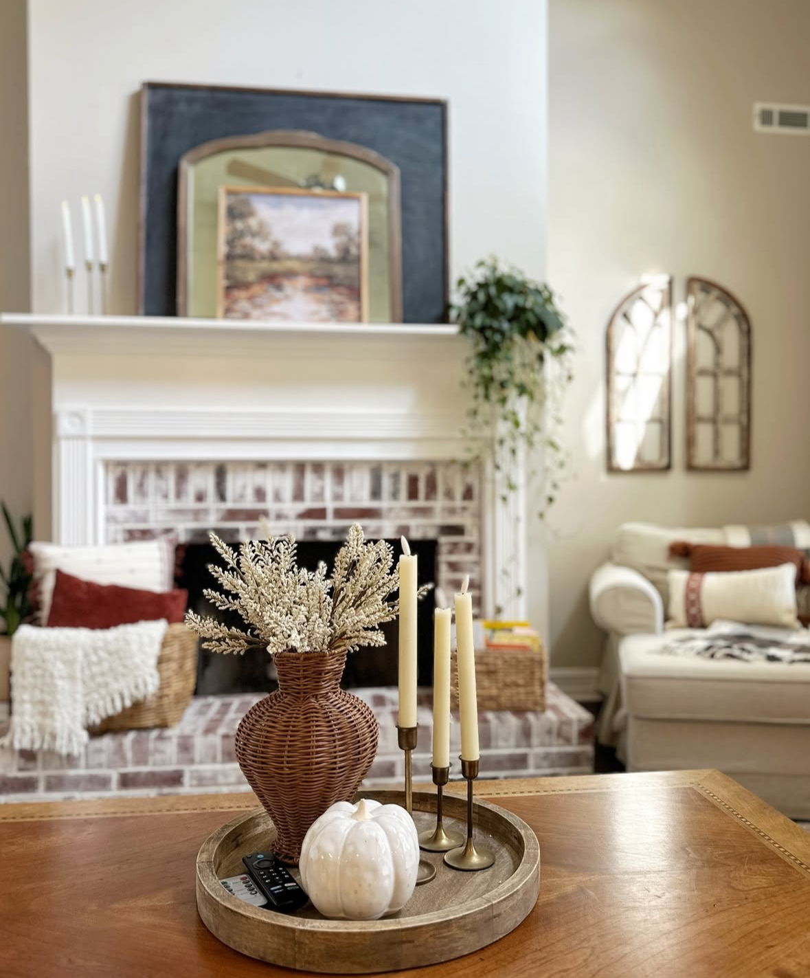 Round wood tray on coffee table with rattan vase of dried stems, three brass candlesticks, and a glossy white pumpkin; brick fireplace backdrop.