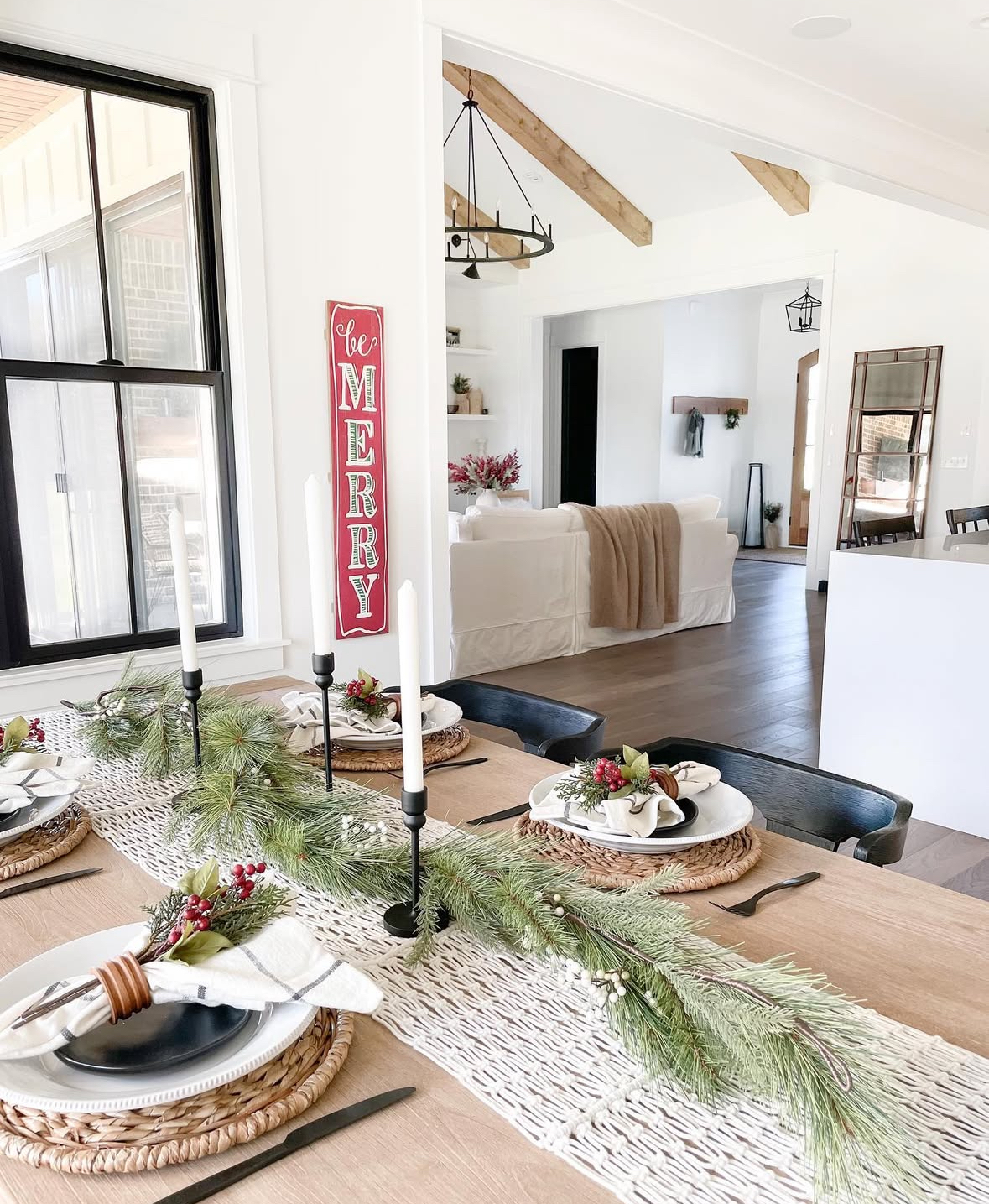 Minimal table of woven chargers, white plates, slim black candleholders, and a long pine garland on a chunky runner.