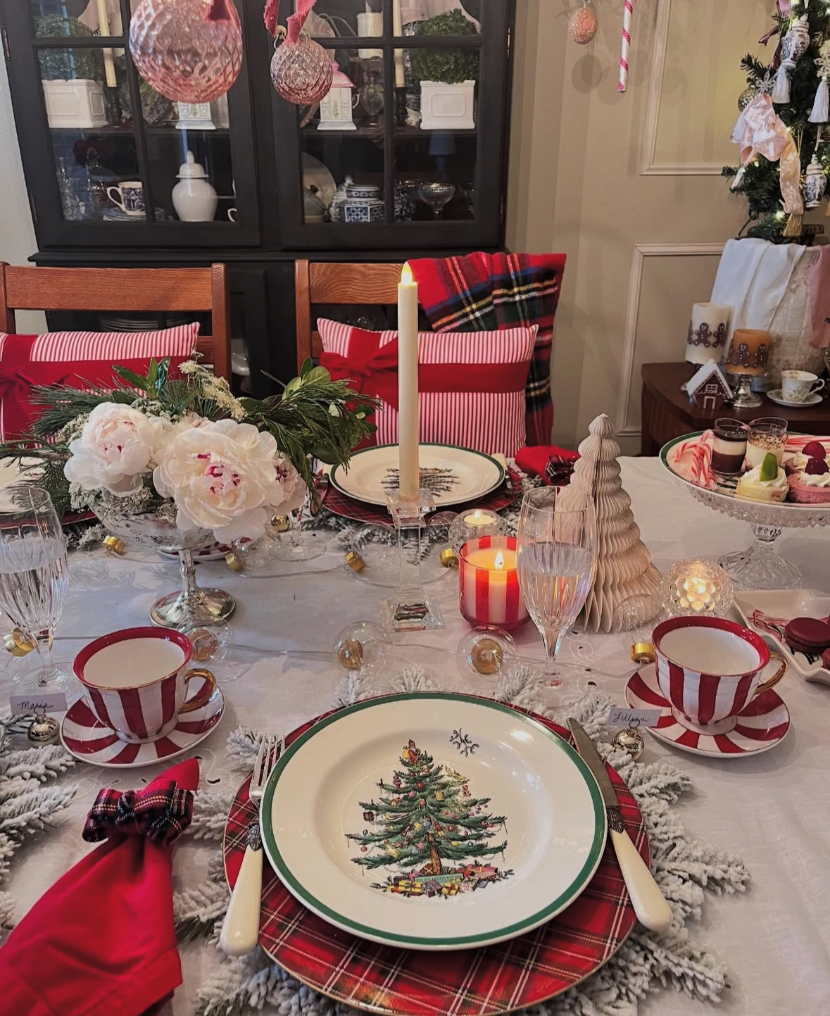 Traditional Christmas tree plates on red plaid chargers with striped teacups, a single taper candle, and festive pillows on chairs.