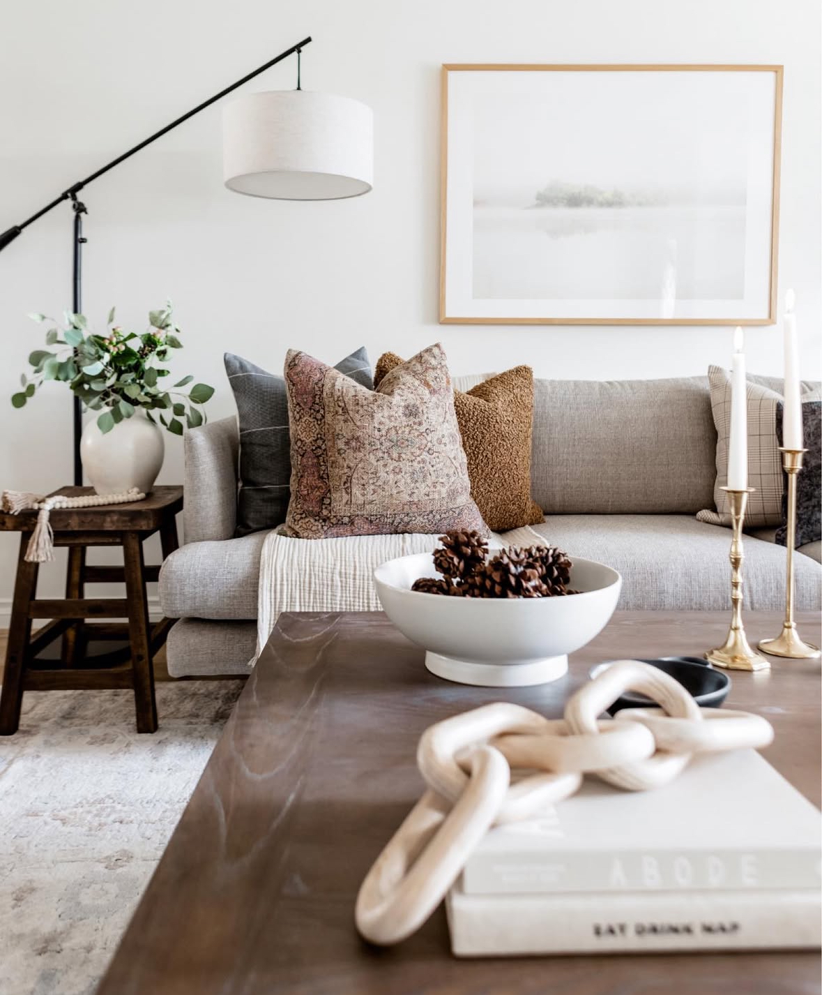 Minimal neutral space with wooden coffee table, white bowl of pinecones, brass candlesticks, wooden chain on books, and eucalyptus in a ceramic vase.