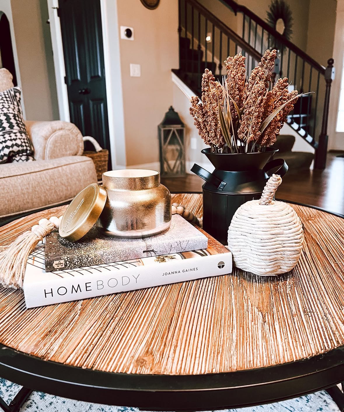 Small round table with stacked books, gold candle jar, black vase of dried grain, and a woven rope pumpkin in a warm, rustic room.