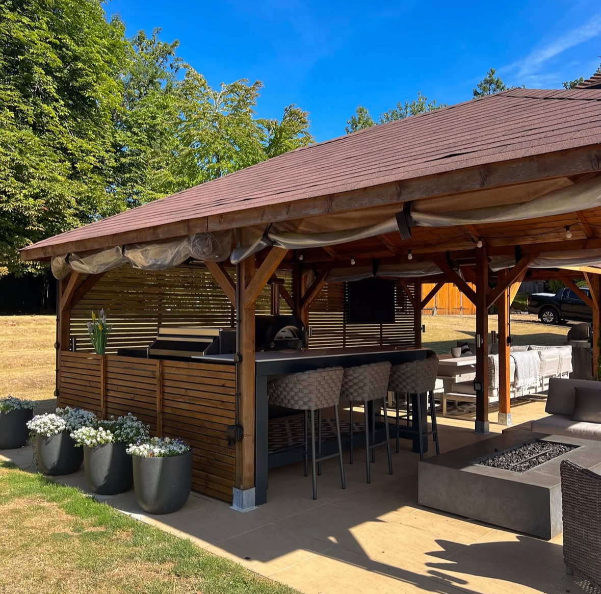 Gabled wood pavilion with bar seating, grill station, and adjacent firepit providing inviting backyard covered outdoor kitchen ideas