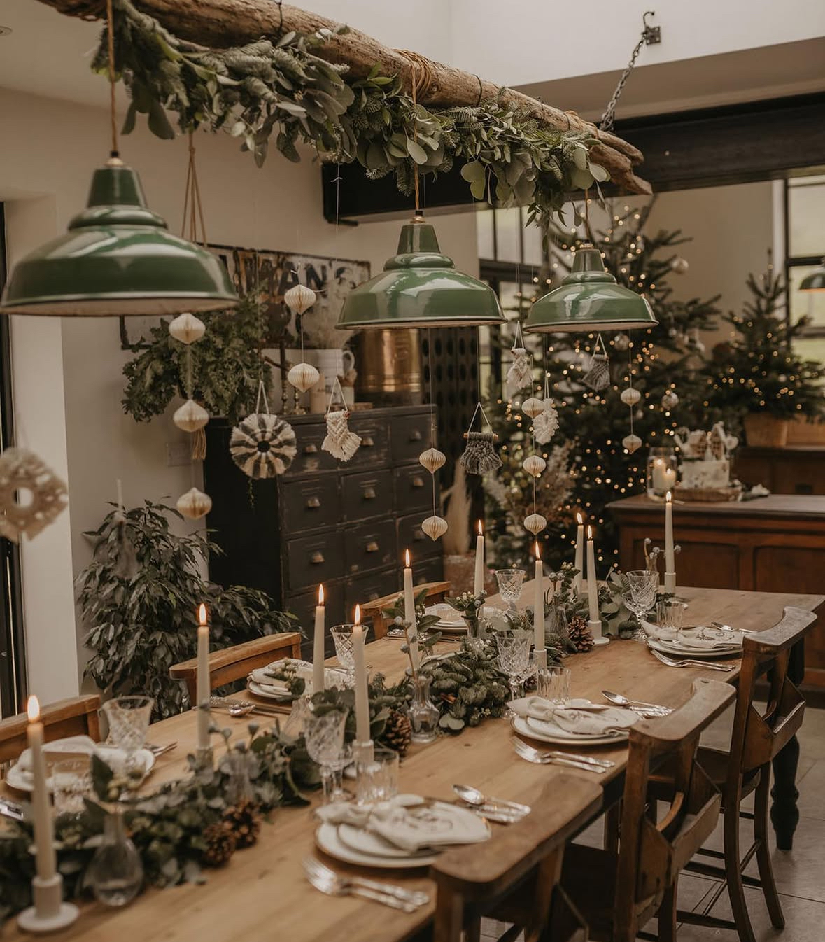 Rustic wood table beneath a suspended log draped in greenery and ornaments, with taper candles and natural foliage.